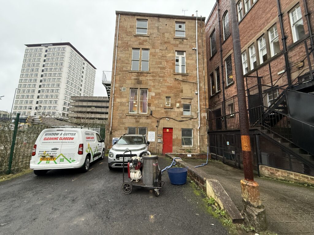 Old brick building with a red door in an alleyway between modern structures.
